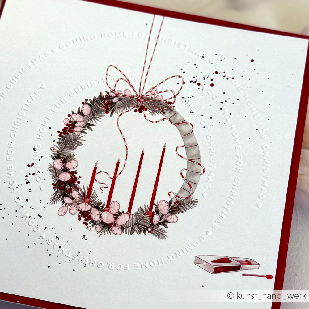 Eine festliche Karte mit dem Schneide- und Prägefolder von Alexandra Renke zeigt einen Kranz, rote Kerzen, ein Band und eine Streichholzschachtel mit dem Schriftzug "Home for Christmas" rund um den Kranz.