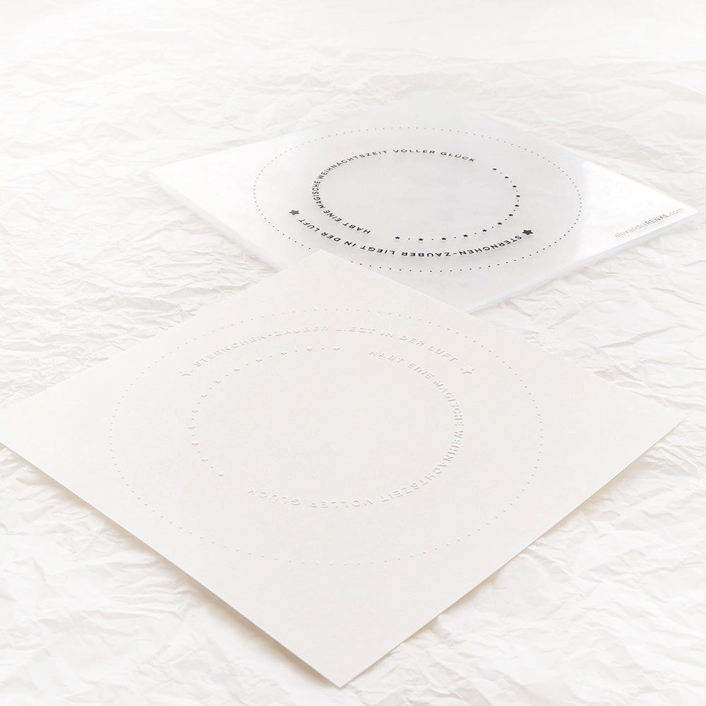 Zwei quadratische weiße Blätter mit perforierten Kreisen und kleinem Text, die auf einer zerknitterten weißen Oberfläche platziert sind, zeigen die komplizierten Details, die mit einem Alexandra Renke Cut & Emboss Folder möglich sind.