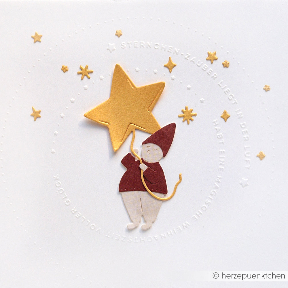 Eine Scherenschnittfigur mit rotem Hut hält einen großen goldenen Stern, umgeben von kleineren Sternen auf weißem Grund - die perfekte Inspiration für Ihre nächste Kreation mit Alexandra Renke Cut & Emboss Folder oder Stanzsystemen.