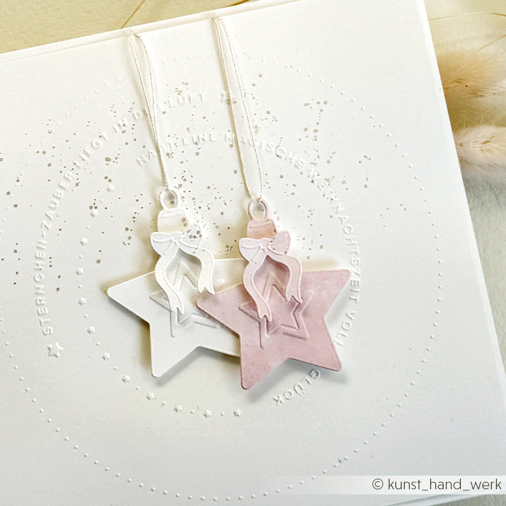 Zwei sternförmige Ornamente - ein weißes und ein rosafarbenes - mit Schleifenband, hergestellt mit dem Alexandra Renke Prägefolder, auf einem strukturierten weißen Hintergrund.