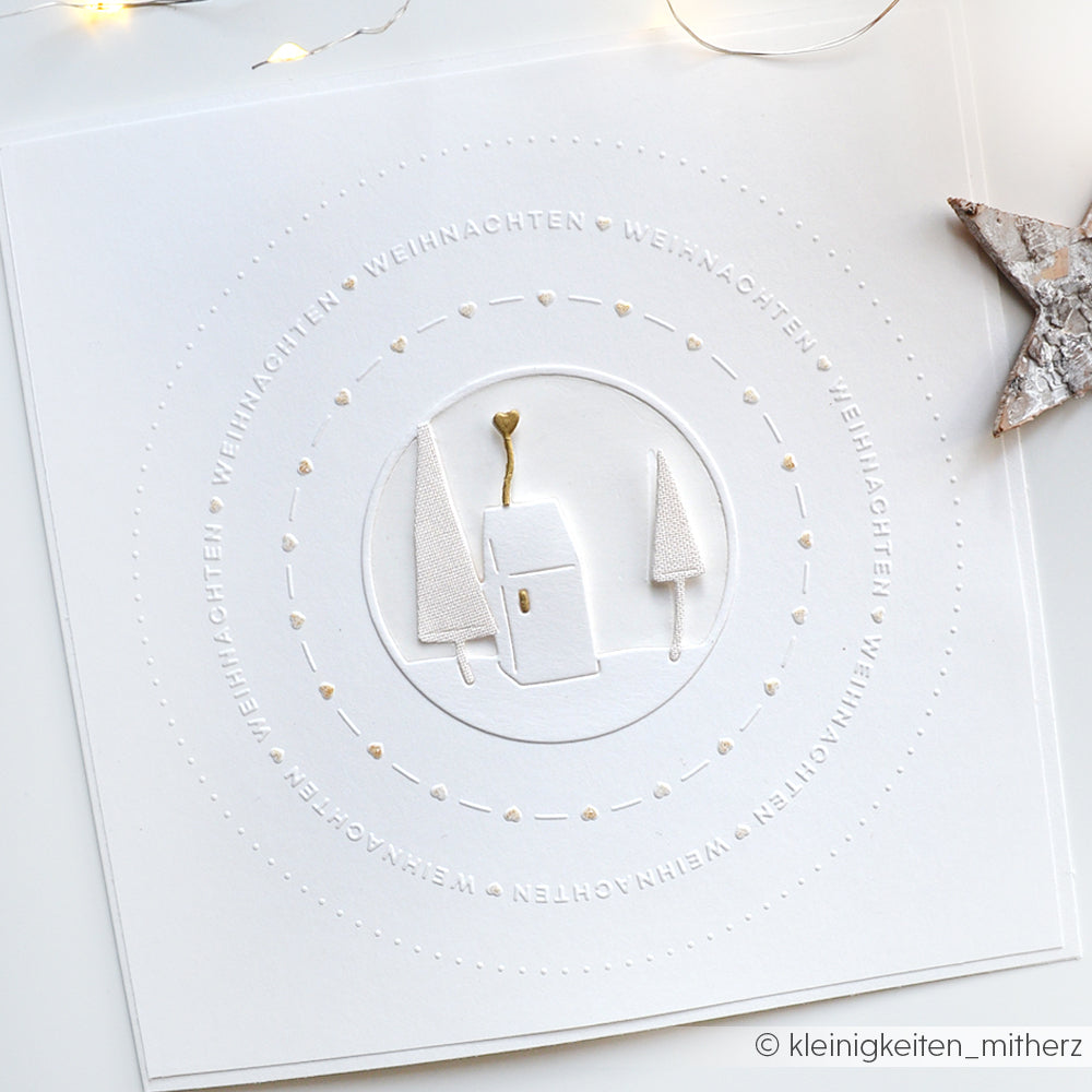 Minimalistische weiße Weihnachtskarte von Alexandra Renke mit einem Haus und Bäumen, die mit dem Cut & Emboss Folder gestaltet wurde. Mit von Typo Kreis geprägten WEIHNACHTEN Textkreisen und eleganten Goldakzenten.