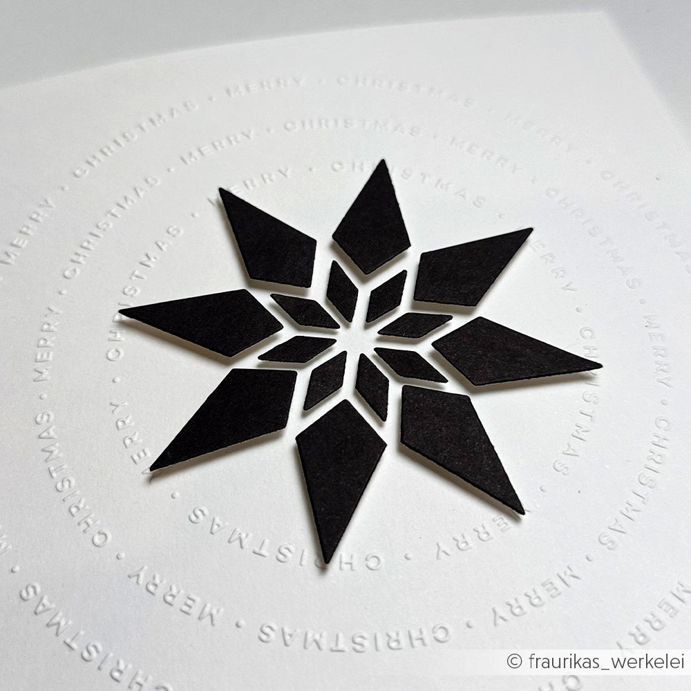 Ein schwarzer, geometrischer Stern auf weißem Papier, der mit dem Alexandra Renke Cut & Emboss Folder hergestellt wurde, wird von einem kreisförmig geprägten Typo Kreis Merry Christmas Text umgeben.