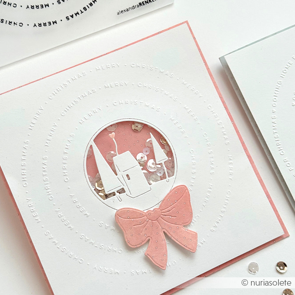 Eine handgefertigte Weihnachtskarte von Alexandra Renke mit einer rosafarbenen Schleife, einer weißen Winterdorfszene, die mit einem Embossing Folder geprägt wurde, und Pailletten in der Mitte.