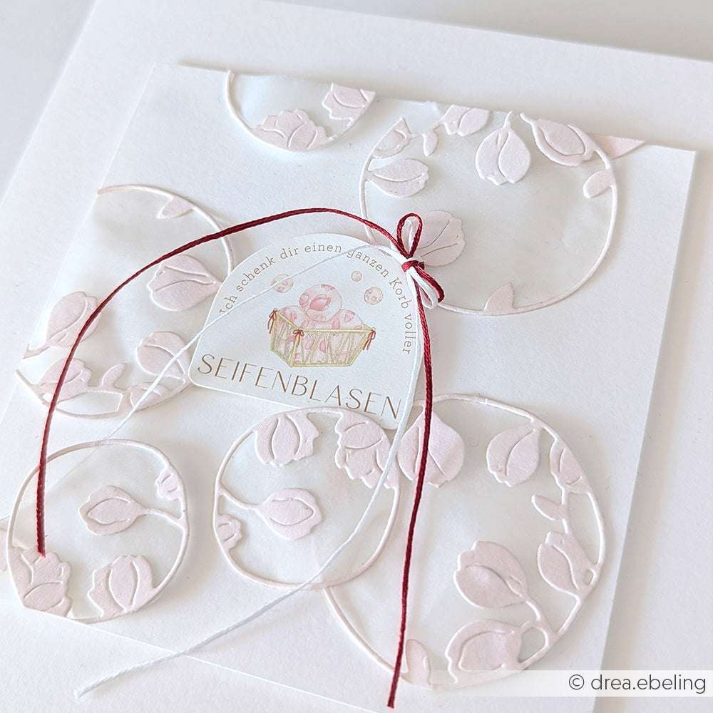 Elegante weiße Karte von Alexandra Renke mit geprägten Blumenkreisen, roten und weißen Schnüren und einem Seifenblasen-Etikett - ideal für Papertie-Projekte oder als einzigartige Ergänzung zu Ihrer Ephemera-Sammlung.