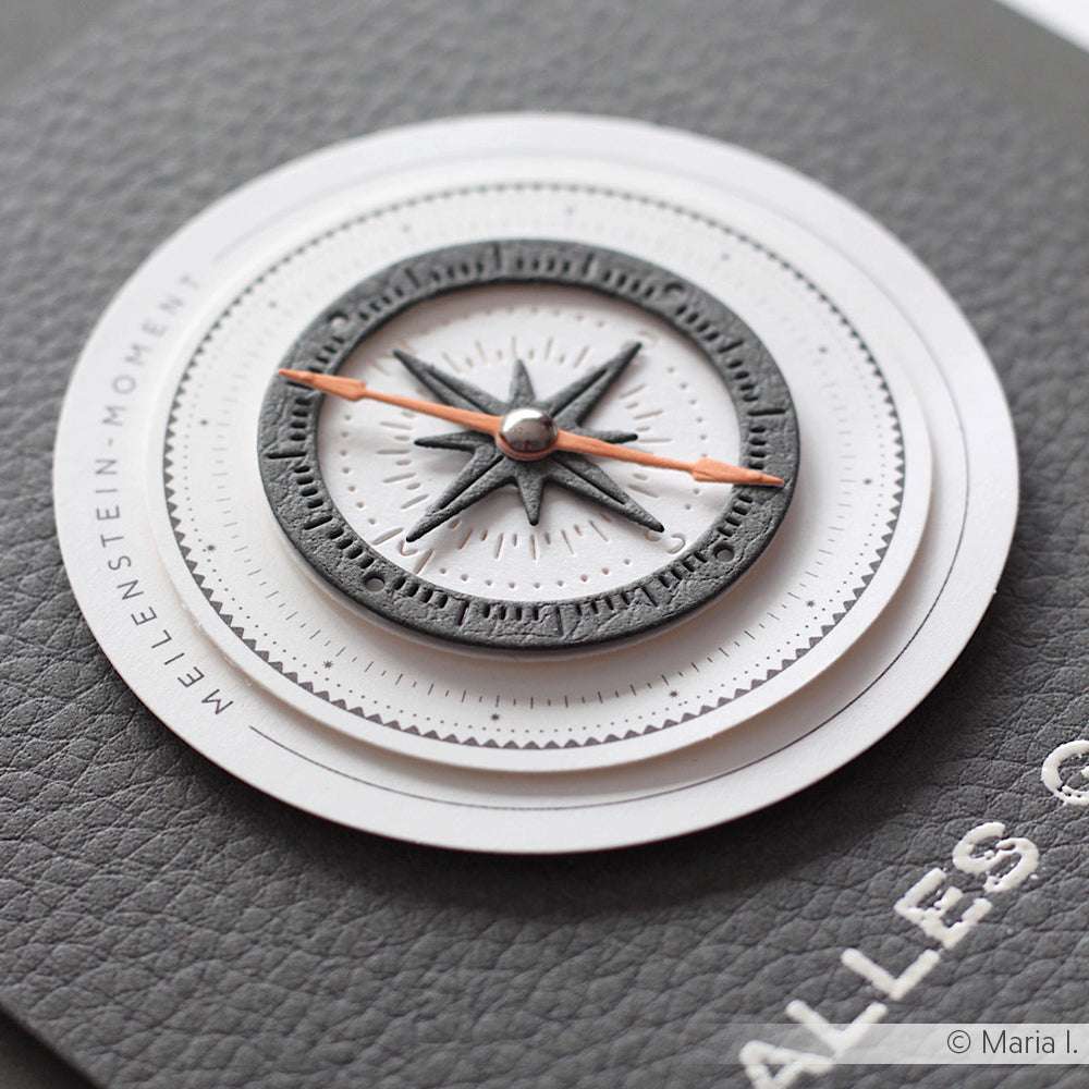 Nahaufnahme eines grau-weißen Kompassmotivs mit orangefarbenen Zeigern auf dunkelgrauem, strukturiertem Hintergrund von Alexandra Renke - ideal für Papertie-Projekte oder zur Ergänzung Ihrer Papierelemente-Sammlung.