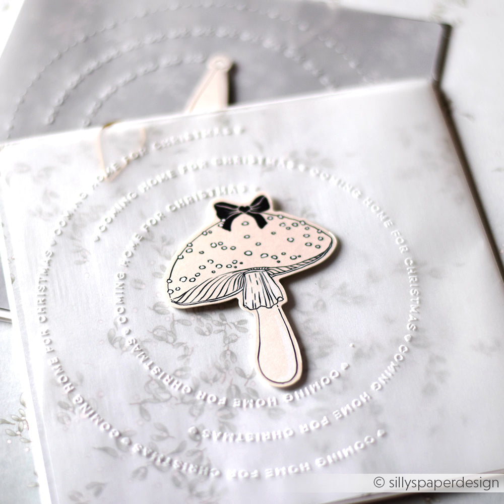 Eine handgefertigte Karte von Alexandra Renke mit Papierelementen, einer Pilzillustration und geprägtem kreisförmigem Text auf einer durchscheinenden Schicht.