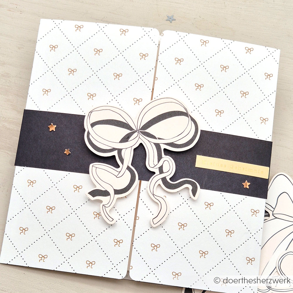 Grußkarte von Alexandra Renke mit einer großen Schleife, kleinen goldenen Sternen, winzigem Papier mit Schleifenmuster, Ephemera-Details und einem schwarzen Band-Ideal für kreative Papertie-Projekte.