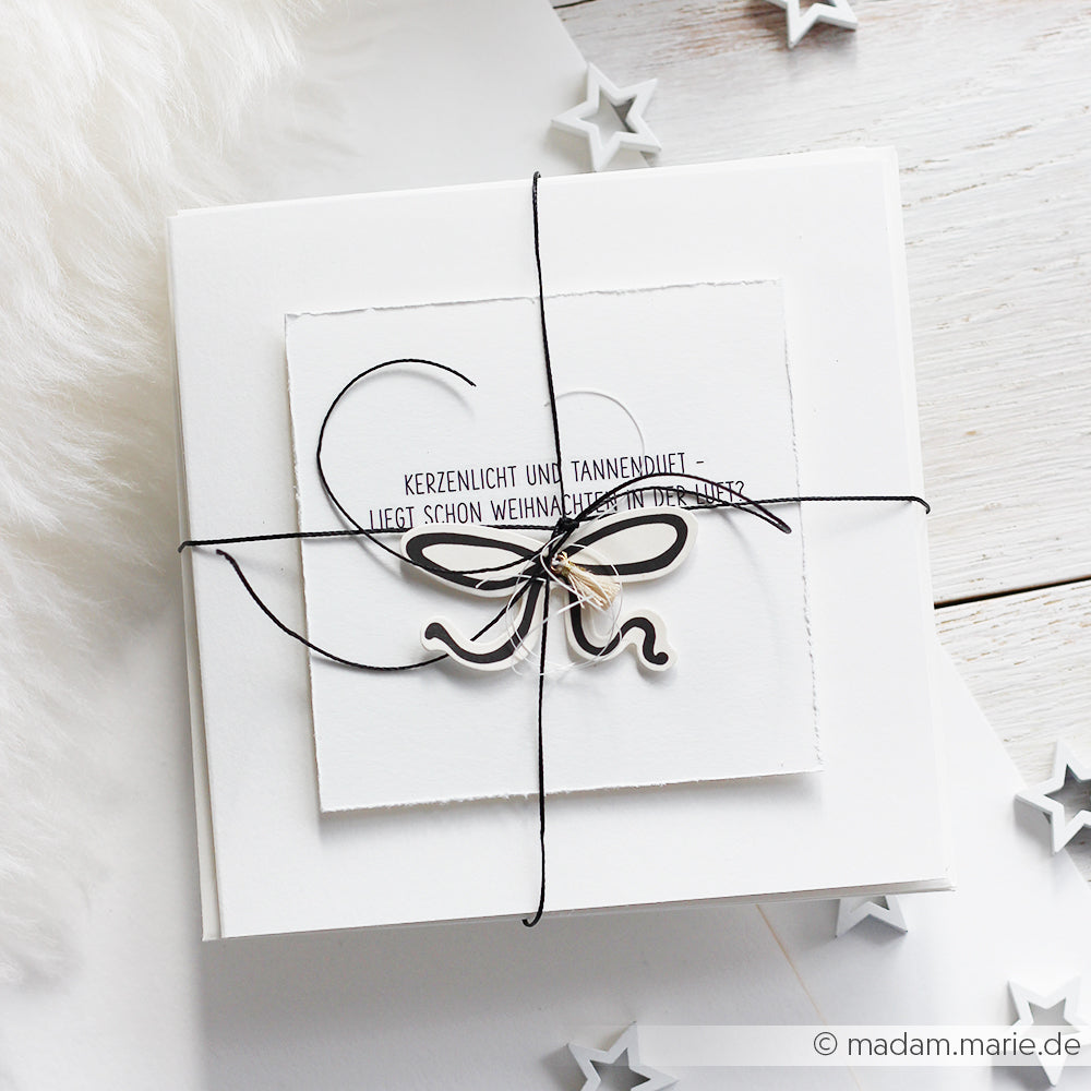Eine weiße Alexandra Renke-Geschenkbox mit Herz- und Schleifenmotiv, verschnürt mit schwarzer Kordel, ruht auf einer weißen Fläche mit Sterndekoration - ideal für Papertie-Projekte oder um kreative Geschenke mit besonderen Ephemera zu versehen.