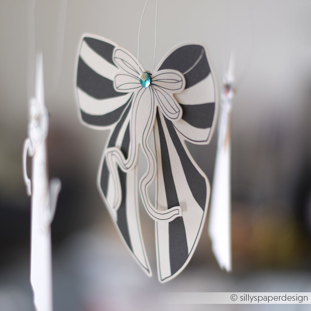 Eine elegante schwarz-weiß gestreifte Papierschleife von Alexandra Renke mit einem blauen Edelstein in der Mitte hängt an einer Schnur und ist als charmante Ephemera gestaltet.