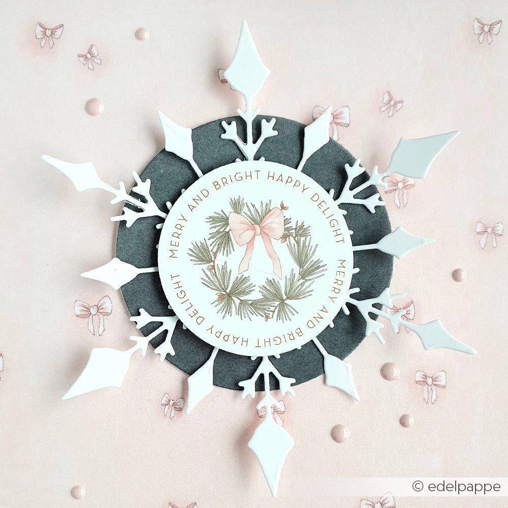 Eine Schneeflocken-Papierkarte von Alexandra Renke mit rosa Schleife, grüner Mitte und dem Text Merry and Bright" - perfekt, um Ihren Papercraft-Projekten festlichen Ephemera-Charme zu verleihen.