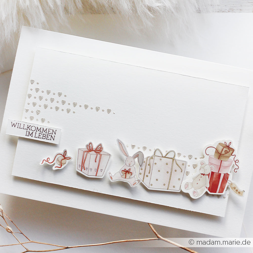 Grußkarte von Alexandra Renke mit Geschenken, Hasen, einer Maus, kleinen Herzen und dem Text "Willkommen im Leben", verziert mit bezaubernden Papercraft-Elementen und entzückenden Ephemera für einen skurrilen Touch.