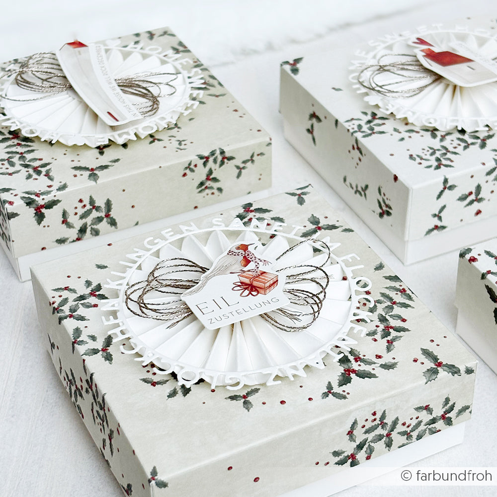 Alexandra Renke dekorative Geschenkboxen mit Stechpalmenmustern, festlichen Anhängern, charmanten Papierelementen und Ephemera werden auf einer weißen Fläche arrangiert.