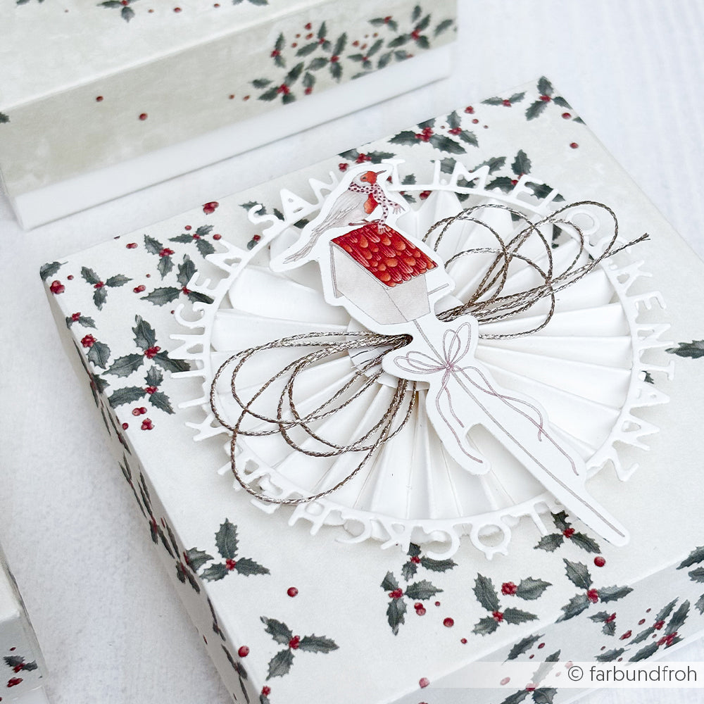 Die Geschenkschachtel von Alexandra Renke ist mit einem Stechpalmenmuster versehen und mit einer Ballerina aus Papierelementen, einer Schleife aus Garn und einer roten Weihnachtsmannmütze verziert.