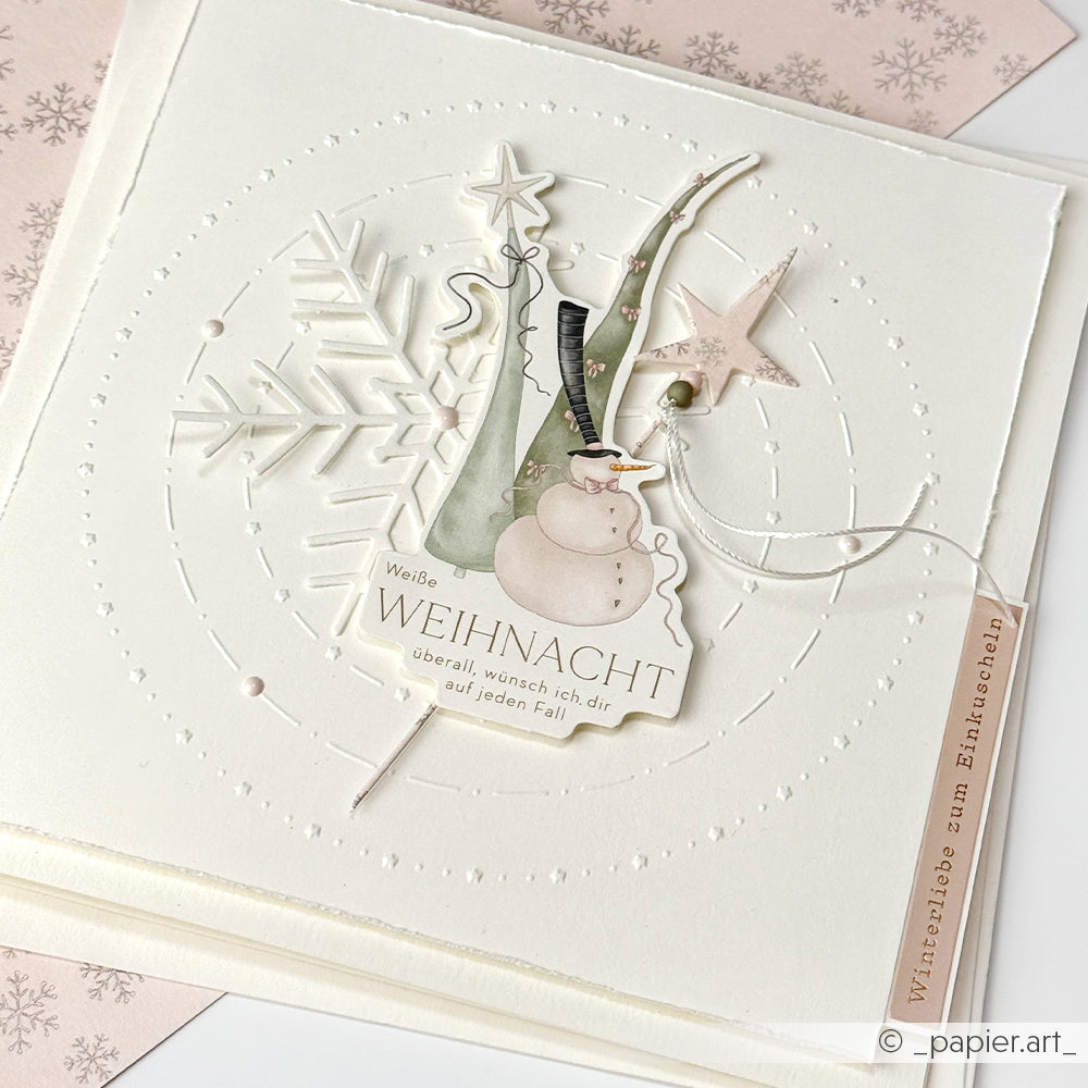Handgefertigte Weihnachtskarte von Alexandra Renke mit zarten Schneeflocken-, Schneemann- und Sternelementen aus Papier und einem deutschen Text auf weißem Papier.