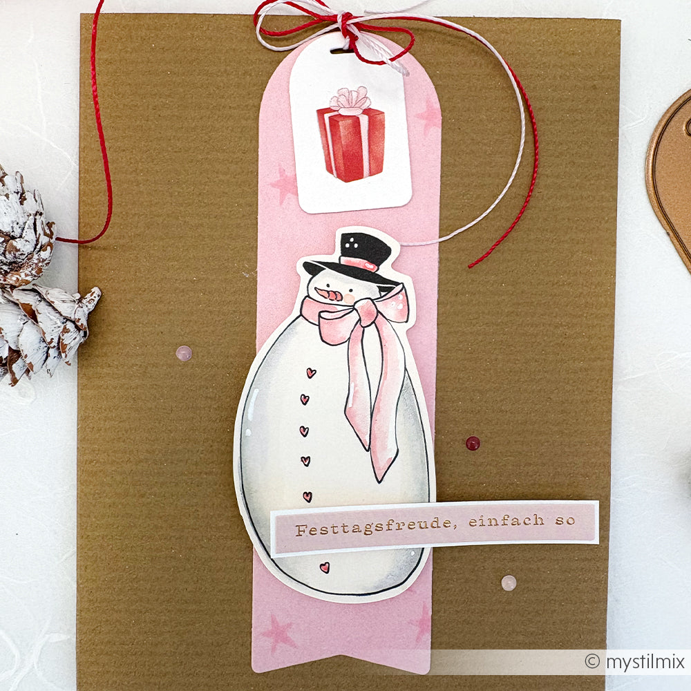 Handgemachte Karte von Alexandra Renke mit Papierelementen: Schneemann, rosa Anhänger und rote Geschenkbox. Mit deutschem Text "Festtagsfreude, einfach so." Ideal für einzigartige Papertie-Projekte und zum Verschenken von Festtagsfreude.