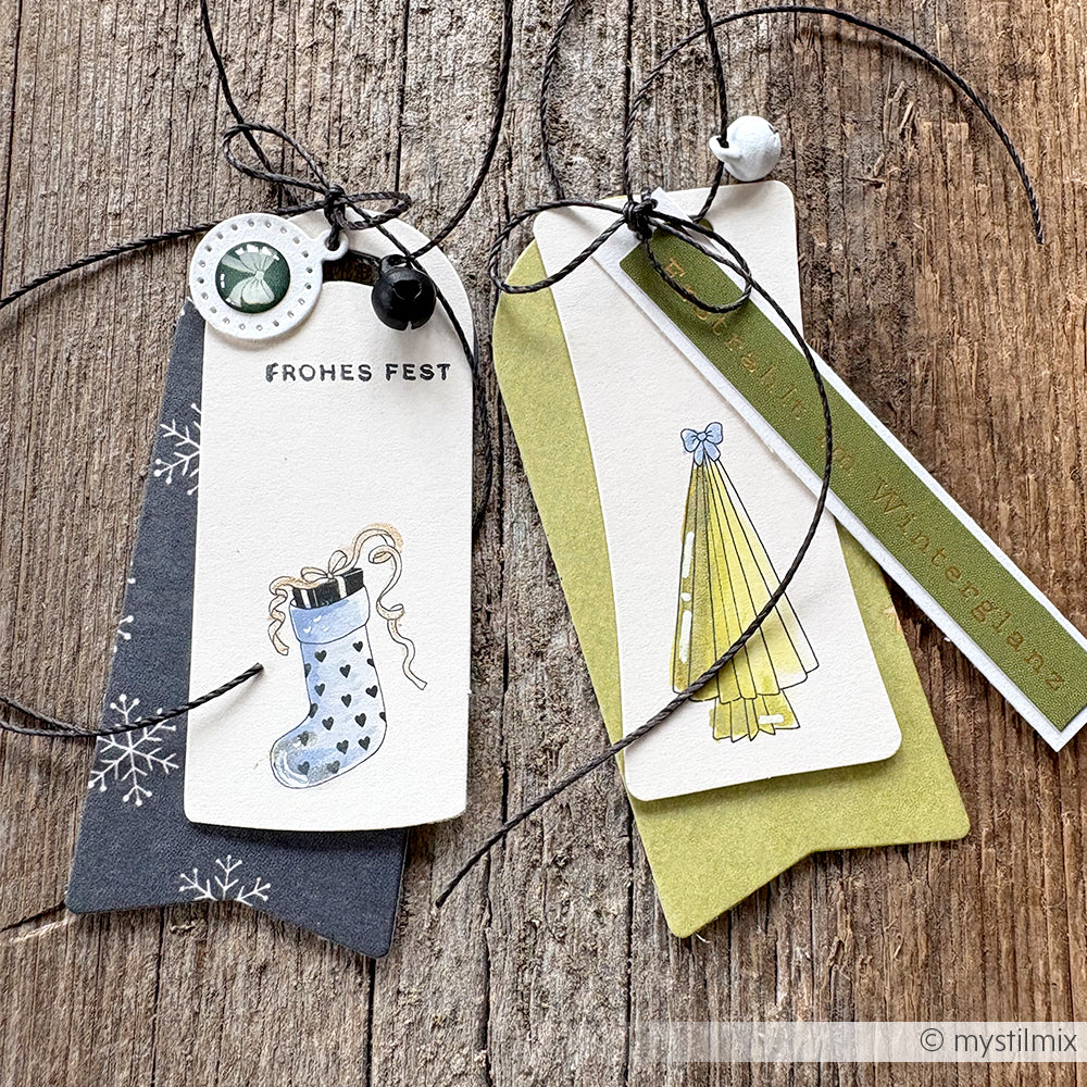 Zwei handgefertigte Geschenkanhänger mit festlichen Motiven und Bändern von Alexandra Renke ruhen auf einer rustikalen Holzoberfläche - perfekt, um Ihren Bastelprojekten eine einzigartige Note zu verleihen.