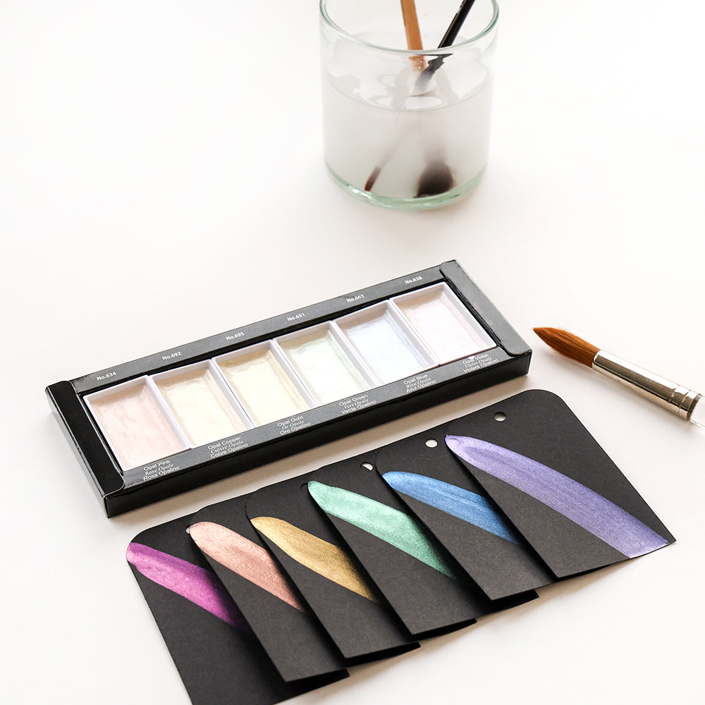 Sechs schillernde Opal Colors Aquarellfarben von Kuretake Gansai Tambi mit Farbmustern auf schwarzen Karten, Aquarellpinseln und einem Glas Wasser auf einem weißen Tisch.