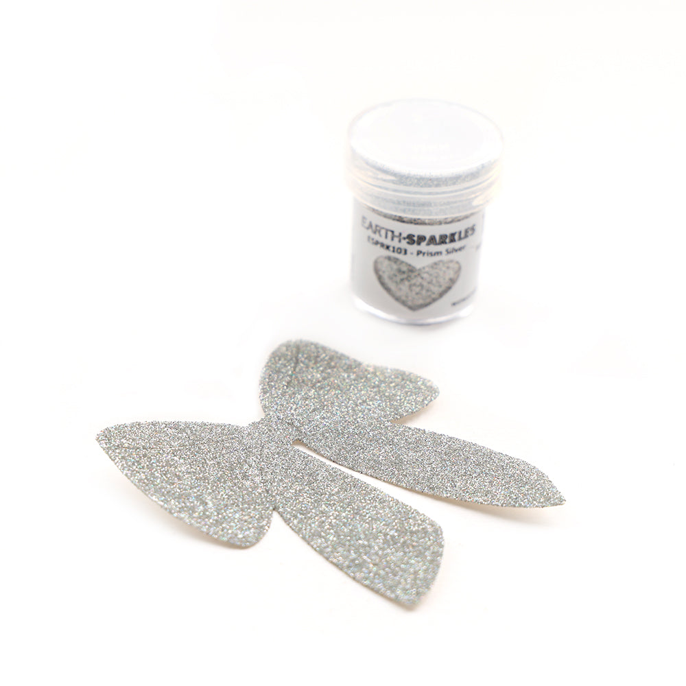 Silberne Glitzerschleifen und ein kleines Döschen WOW Embossing Powder Bio Glitter mit einem herzförmigen Etikett und pflanzlichen Glitzerflocken, dargestellt auf weißem Hintergrund.