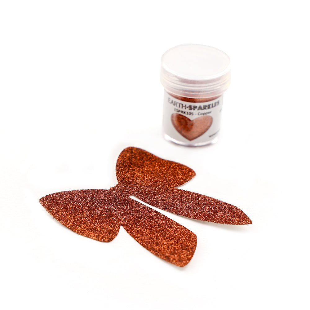 Hinter zwei kupferfarbenen Glitzerschleifen auf weißem Hintergrund befindet sich eine kleine Dose WOW Embossing Powder, die mit pflanzlichen Glitzerflocken für einen umweltfreundlichen Glanz sorgt.