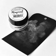 Eine kleine Dose Lindy's Stamp Gang Magical Powder in milchigem Weiß steht neben einem schwarzen Anhänger, der mit dem weißen Pigment bestrichen wurde und dessen einzigartige Farbe hervorhebt.