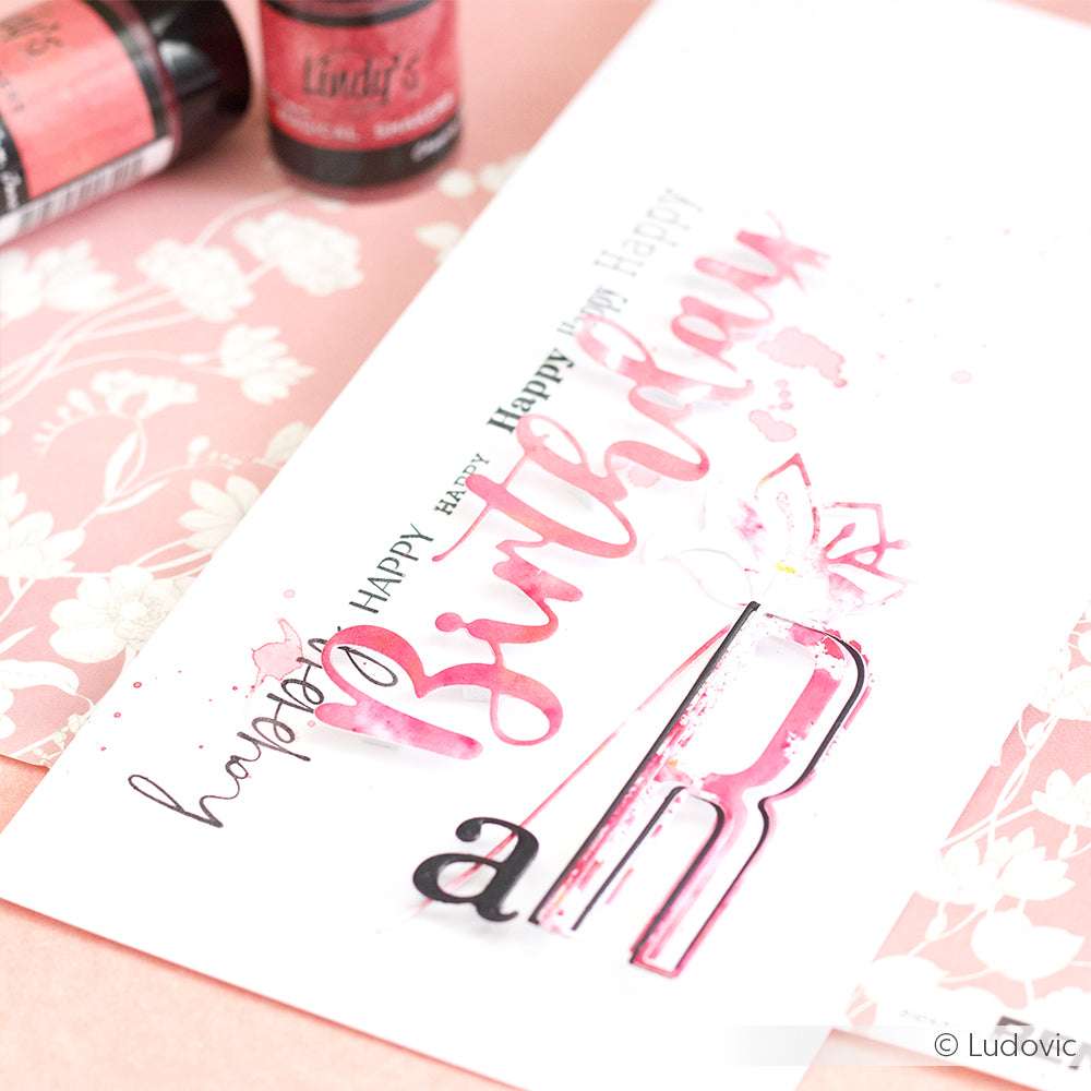 Handgemachte Lindy's Stamp Gang Karte mit "Happy Birthday" in rosa Aquarell auf einem floralen Hintergrund, mit magischem Pulver für lebendige Farbmischungen; inklusive sichtbarem Bastelmaterial.