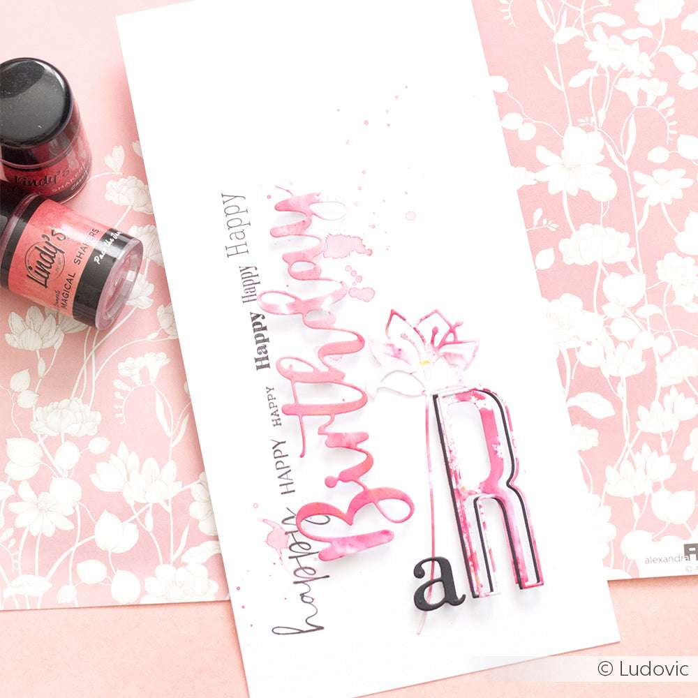 Handgemachte Geburtstagskarte mit rosa und schwarzer Schrift und Farbspritzern, hergestellt mit Lindy's Stamp Gang hochpigmentiertem Pulver, mit dekorativem Kunstzubehör in der Nähe.