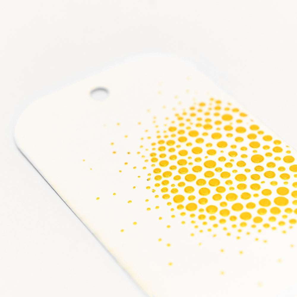 Der weiße Anhänger von Crafters Workshop mit abgerundeten Kanten und einem Loch ist mit einem gelben Rasterpunktmuster versehen - perfekt für Mixed Media-Projekte mit Stencil Butter oder Modellierpaste.