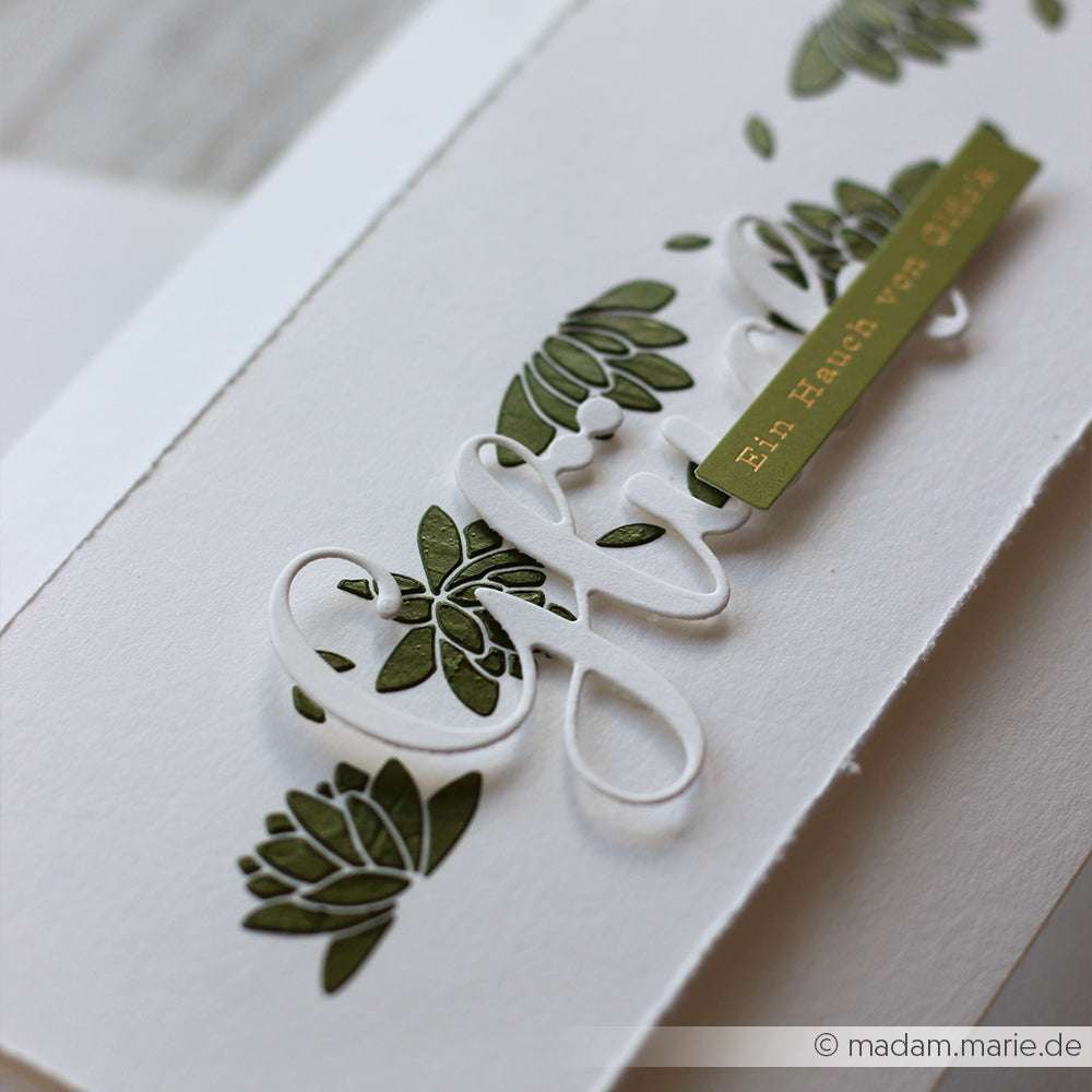 Weiße Karte mit grünem Blumenmuster, erhabener Schrift und einem grünen Etikett mit der Aufschrift "Ein Hauch von Glück". Mixed-Media-Techniken mit The Crafters Workshop Stencil Butter und Modellierpaste sorgen für elegante Textur und Tiefe.