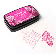 Ein rosafarbenes Tsukineko VersaFine Clair Stempelkissen und zwei weiße Anhänger mit rosa gestempeltem dekorativem Text und Motiven.