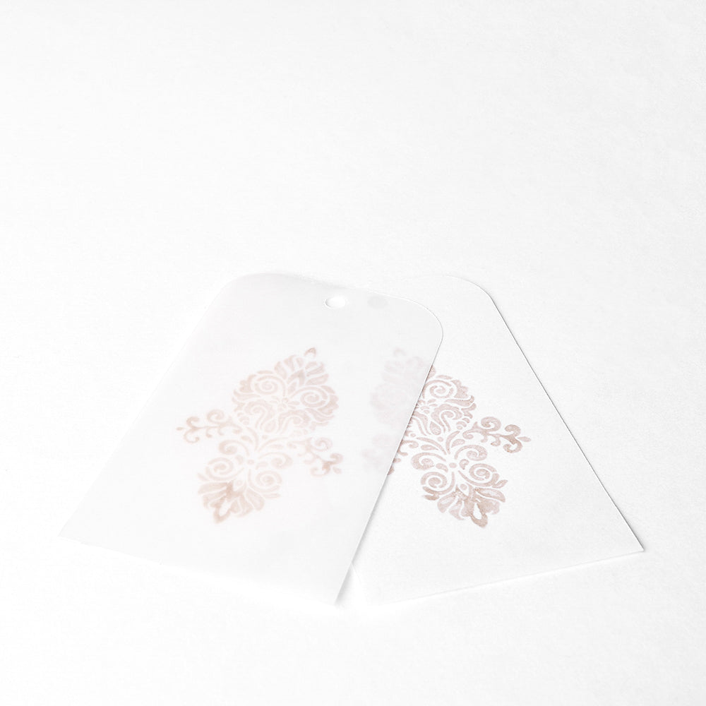 Zwei durchscheinende Anhänger mit verschnörkelten, hellrosa Blumenmustern auf Weiß, die mit Tsukineko Brilliance Stempelkissen für schimmernde Stempeleffekte wunderschön akzentuiert sind.