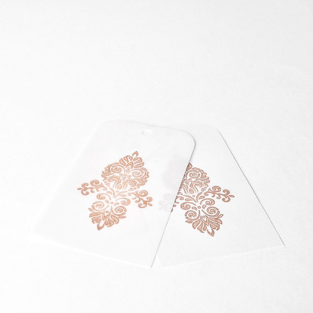 Zwei durchscheinende Anhänger mit verschnörkelten goldenen Motiven, die mit Tsukineko Brilliance Stempelkissen für einen metallischen Schimmer hergestellt wurden, liegen leicht überlappend auf einer weißen Fläche.