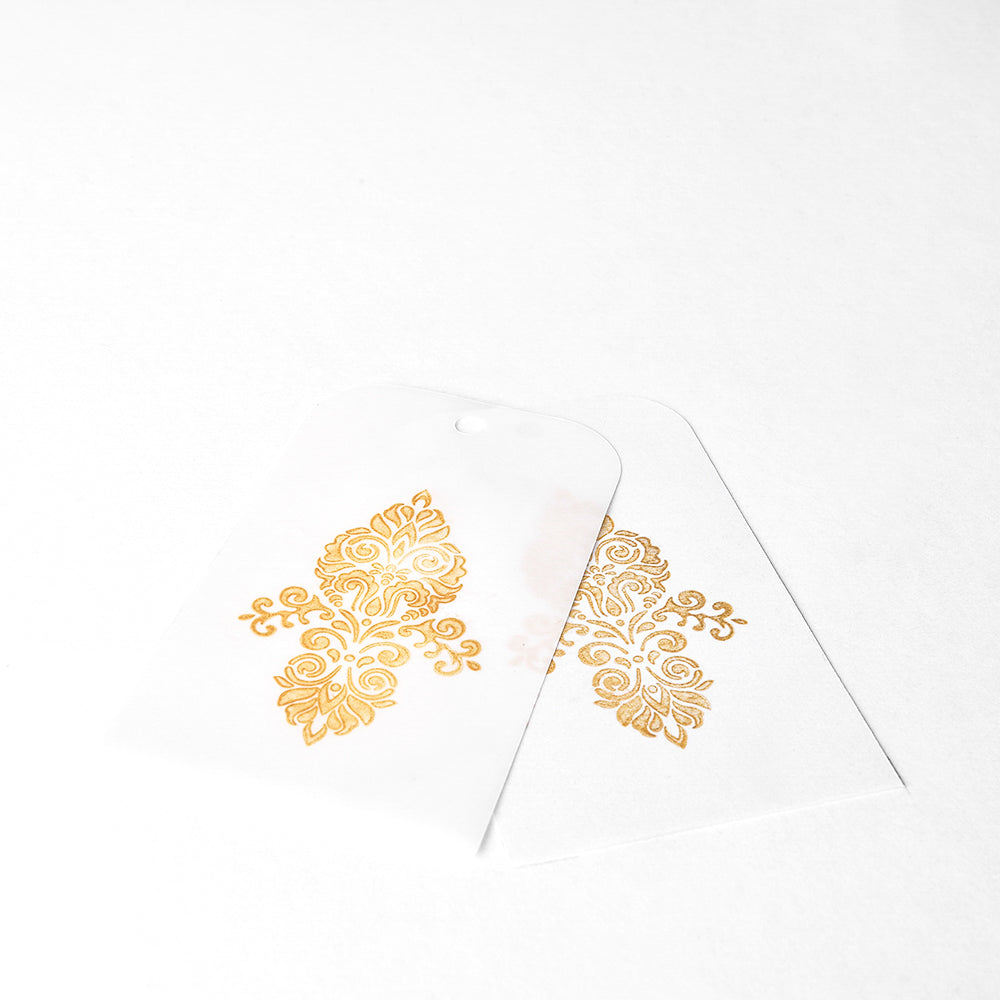 Zwei transparente Geschenkanhänger mit goldfarbenen Blumenmustern, hergestellt mit hochpigmentierter Tinte von Tsukineko Brilliance Stempelkissen, auf weißem Hintergrund.