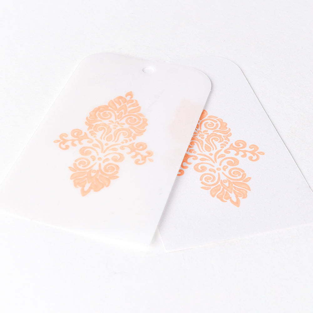 Zwei Karten mit verschnörkelten, pfirsichfarbenen Blumenmustern, die mit Tsukineko Versa Magic Pigment-Tinte Stempelkissen erstellt wurden, überlappen sich auf einem weißen Hintergrund.