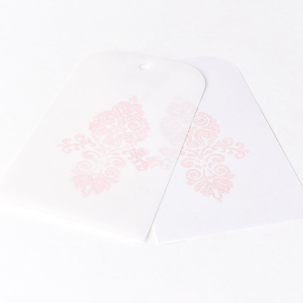 Zwei weiße Anhänger mit zartrosa Blumenmustern, die mit Tsukineko Versa Magic Stempelkissen Pigment-Tinte hergestellt wurden - einer durchscheinend und über den anderen gelegt - werden auf einem weißen Hintergrund angezeigt.