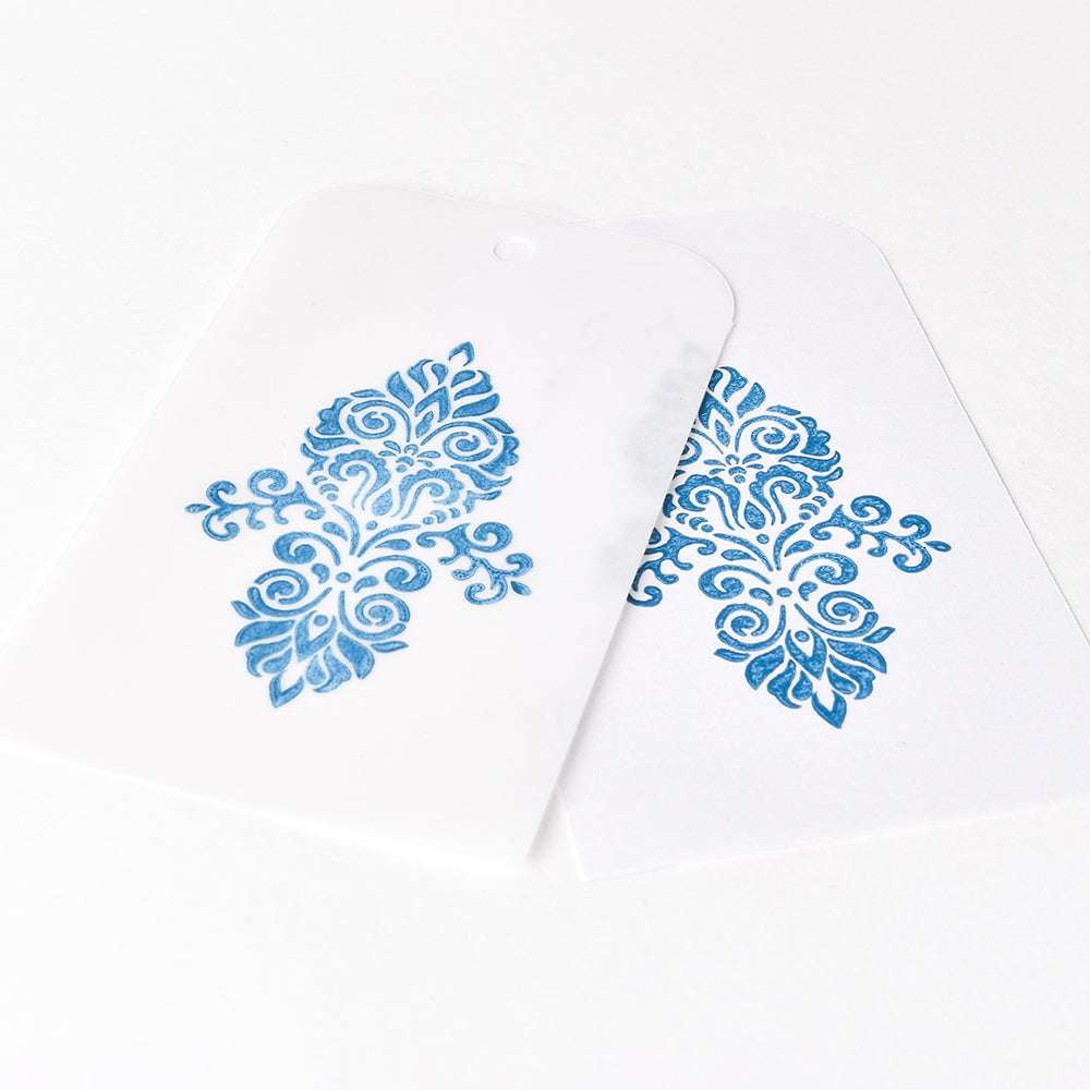 Zwei weiße Anhänger mit verschnörkelten blauen Blumenmustern, hergestellt mit Tsukineko Versa Magic Stempelkissen und wasserbasierter Pigment-Tinte, überlappen sich auf weißem Hintergrund für einzigartige Stempelergebnisse.