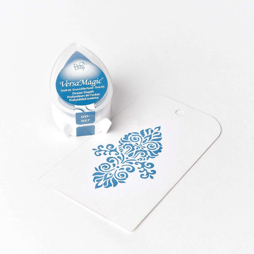 Ein blaues Tsukineko VersaMagic Stempelkissen liegt neben einem weißen Anhänger mit einem blauen, verschnörkelten Stempelmuster und demonstriert ein gestochen scharfes Stempelergebnis mit Pigmenttinte auf Wasserbasis.