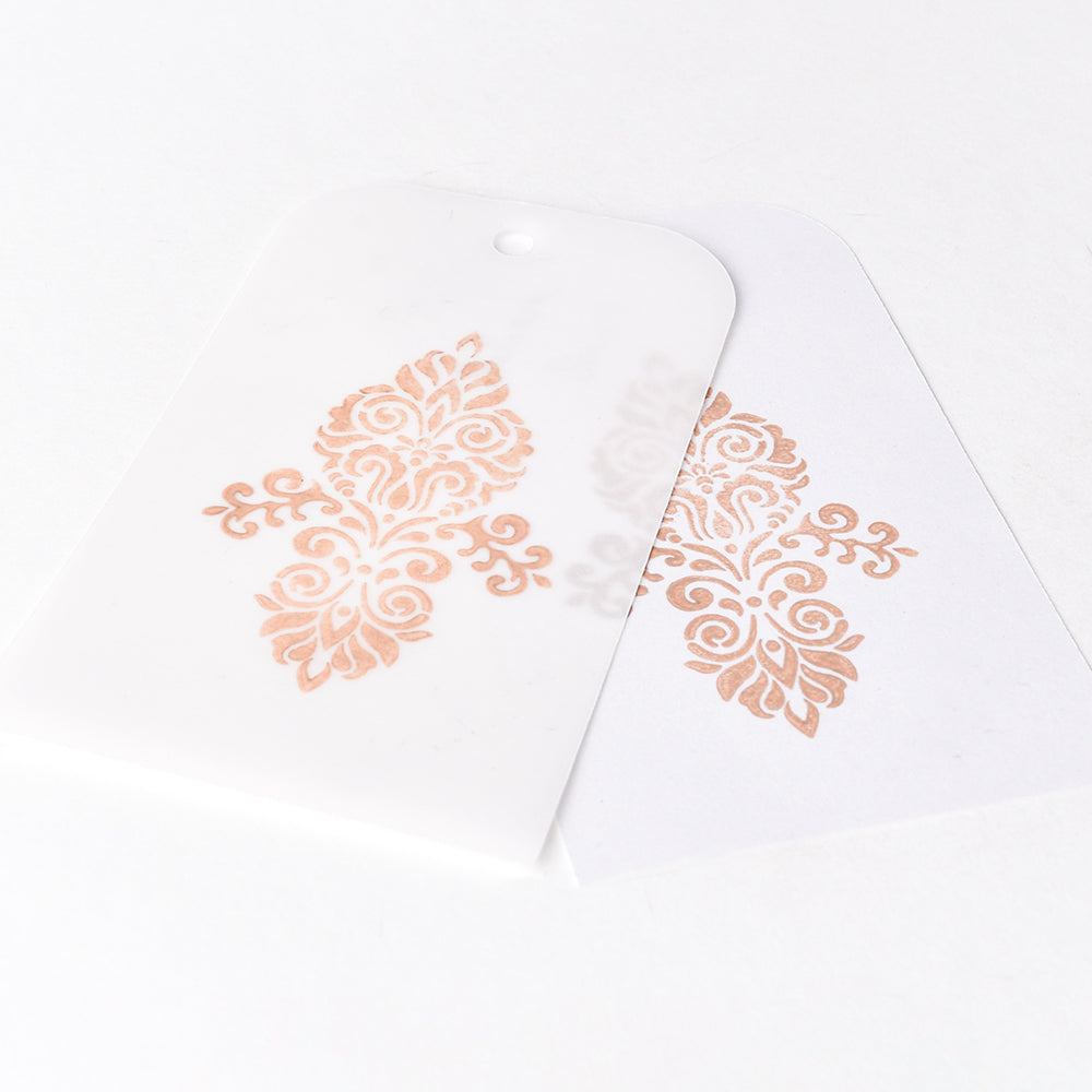 Zwei weiße Tsukineko-Anhänger mit verschnörkelten Goldmustern - einer teilweise transparent und den anderen überlappend - sind ideal für Stempeln auf glattem Papier mit Versa Magic Stempelkissen.
