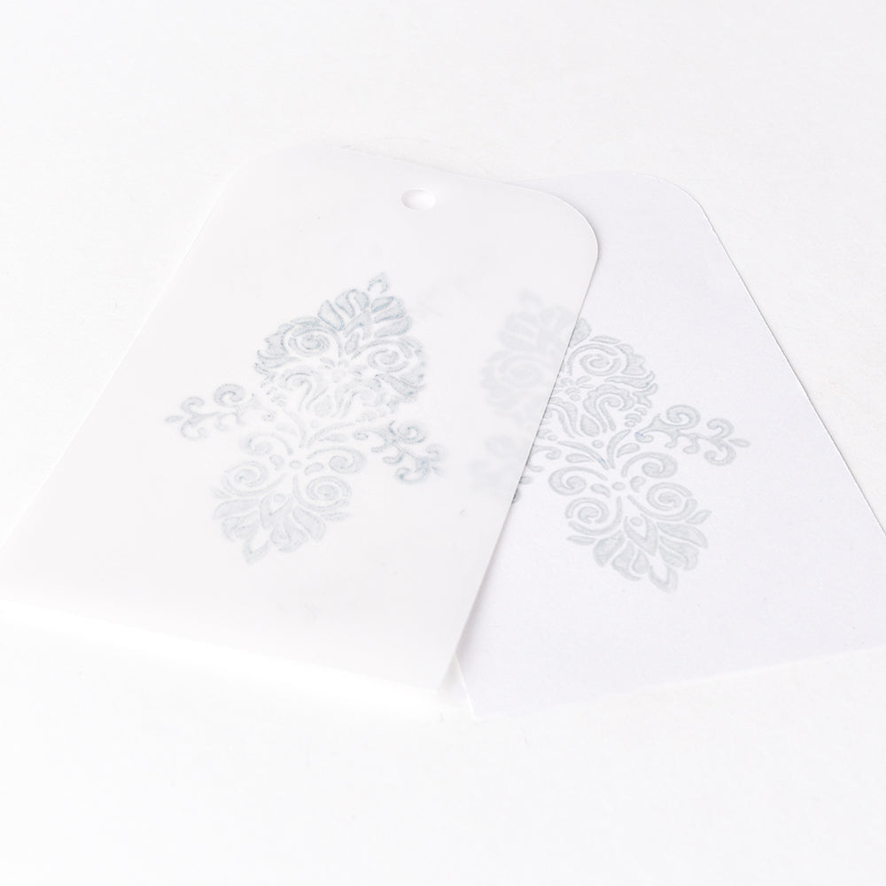 Zwei durchscheinende Karten mit verschnörkelten silbernen Motiven, die sich gegenseitig überlappen, auf weißem Hintergrund - ideal für die Verwendung mit Tsukineko Versa Magic Stempelkissen oder wasserbasierter Pigment-Tinte für gestochen scharfe Ergebnisse auf glattem Papier.