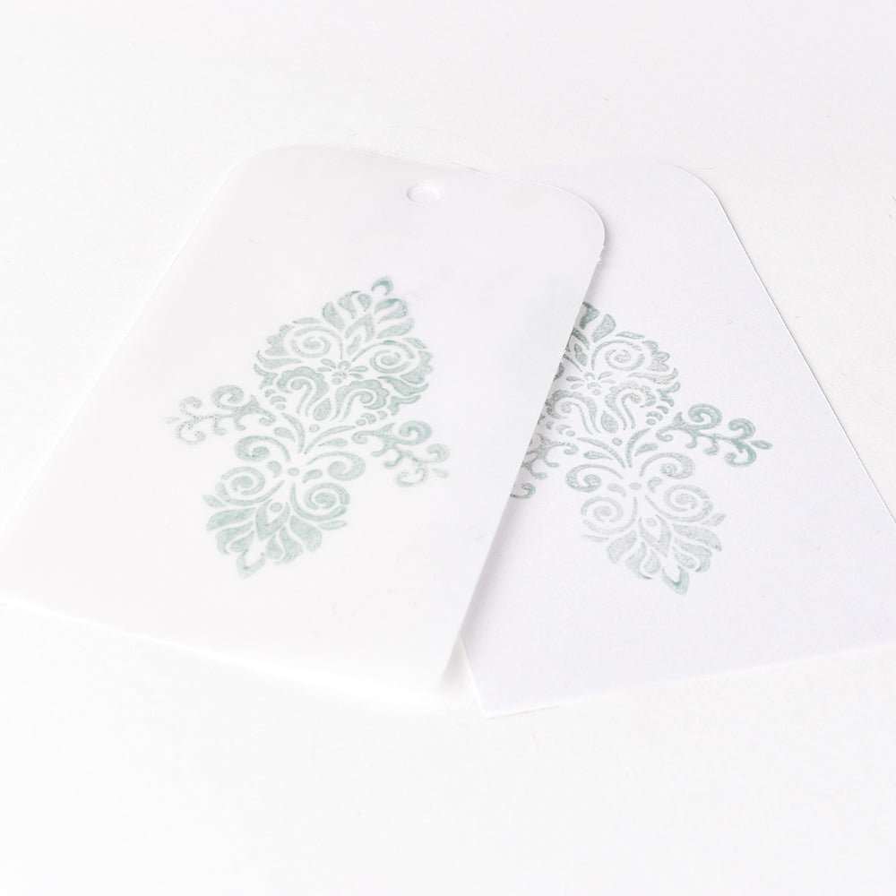 Zwei weiße Anhänger mit verschlungenen hellgrünen Blumenmustern, hergestellt mit Tsukineko Versa Magic Stempelkissen Pigment-Tinte, wobei ein Anhänger den anderen teilweise überlappt.