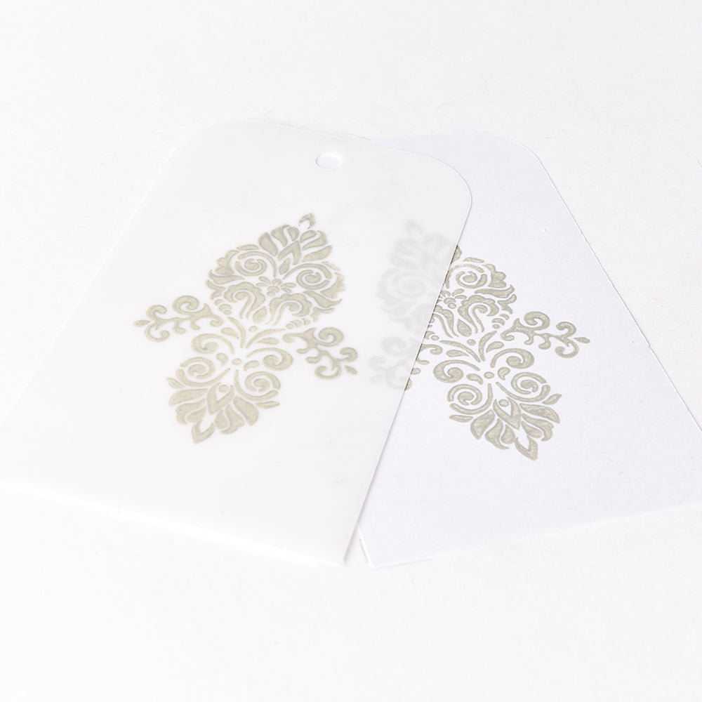 Zwei dekorative Geschenkanhänger - einer transluzent und einer weiß - zeigen goldene Blumenmuster, die mit Tsukineko Pigment-Tinte Stempelkissen auf einer weißen Oberfläche wunderschön hervorgehoben sind.