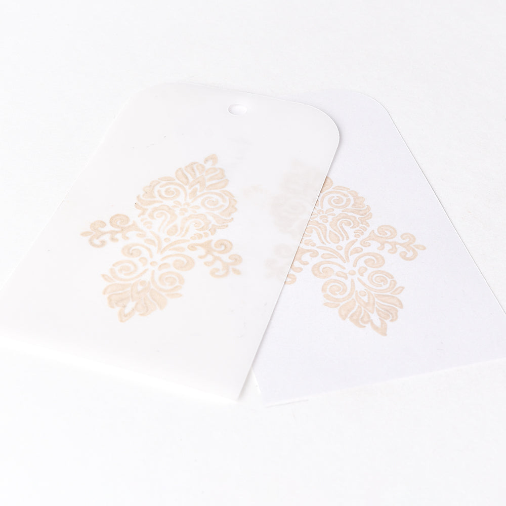 Zwei weiße Anhänger mit verschnörkelten beigefarbenen Blumenmustern - erstellt mit der Tsukineko Versa Magic Stempelkissen Pigment-Tinte - liegen leicht überlappend auf weißem Hintergrund. Perfekt für Stempeln auf glattem Papier.