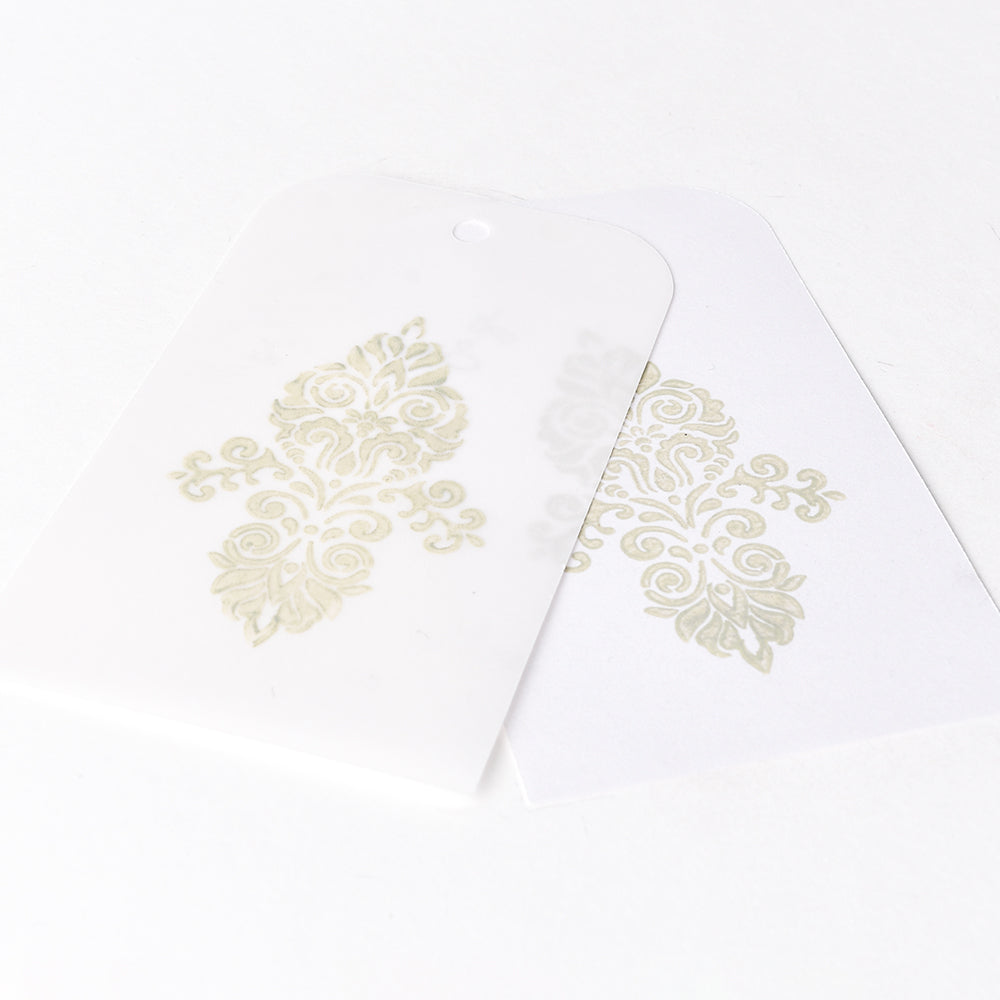 Zwei dekorative weiße Anhänger mit verschnörkelten goldenen Mustern - einer durchscheinend, der andere überlappend - unterstreichen den Charme der wasserbasierten Pigmenttinte, die mit Tsukineko Versa Magic Stempelkissen für einen raffinierten Touch geschaffen wurde.