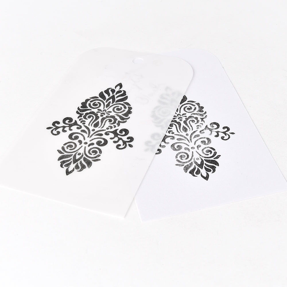 Zwei weiße Anhänger mit verschnörkelten schwarzen Blumenmustern, die sich teilweise überlappen, zeigen beeindruckende Prägeergebnisse, die mit Tsukineko Versa Magic Stempelkissen erstellt wurden.