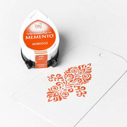 Ein kleines, orangefarbenes Tsukineko Memento Stempelkissen in Marokko liegt neben einem weißen Anhänger mit einem orangefarbenen Blumenmuster, das mit seiner hochpigmentierten Farbe gestempelt ist.