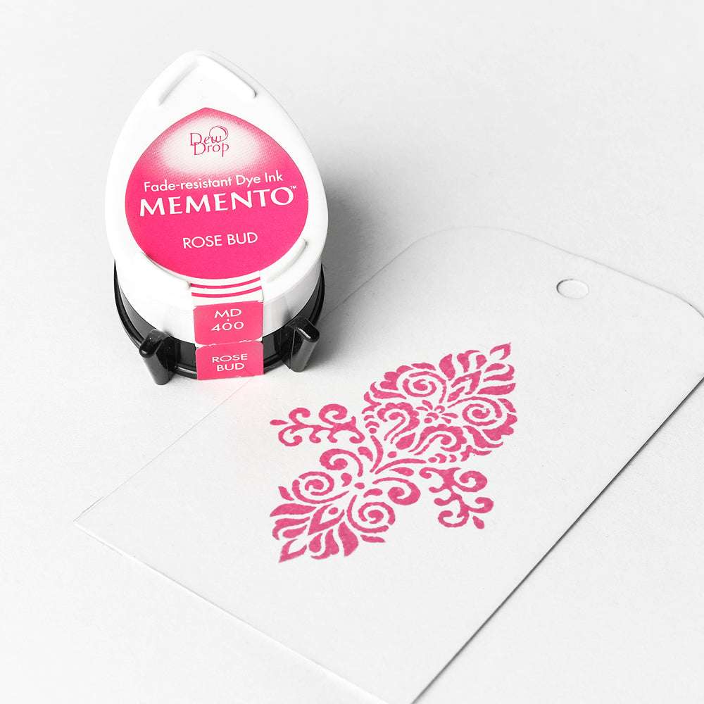 Ein Tsukineko Memento Stempelkissen Rose Bud wird neben einem weißen Anhänger präsentiert, der mit einem rosafarbenen, verschnörkelten Blumenmuster bestempelt ist und dessen hochpigmentierte Farbe hervorhebt.