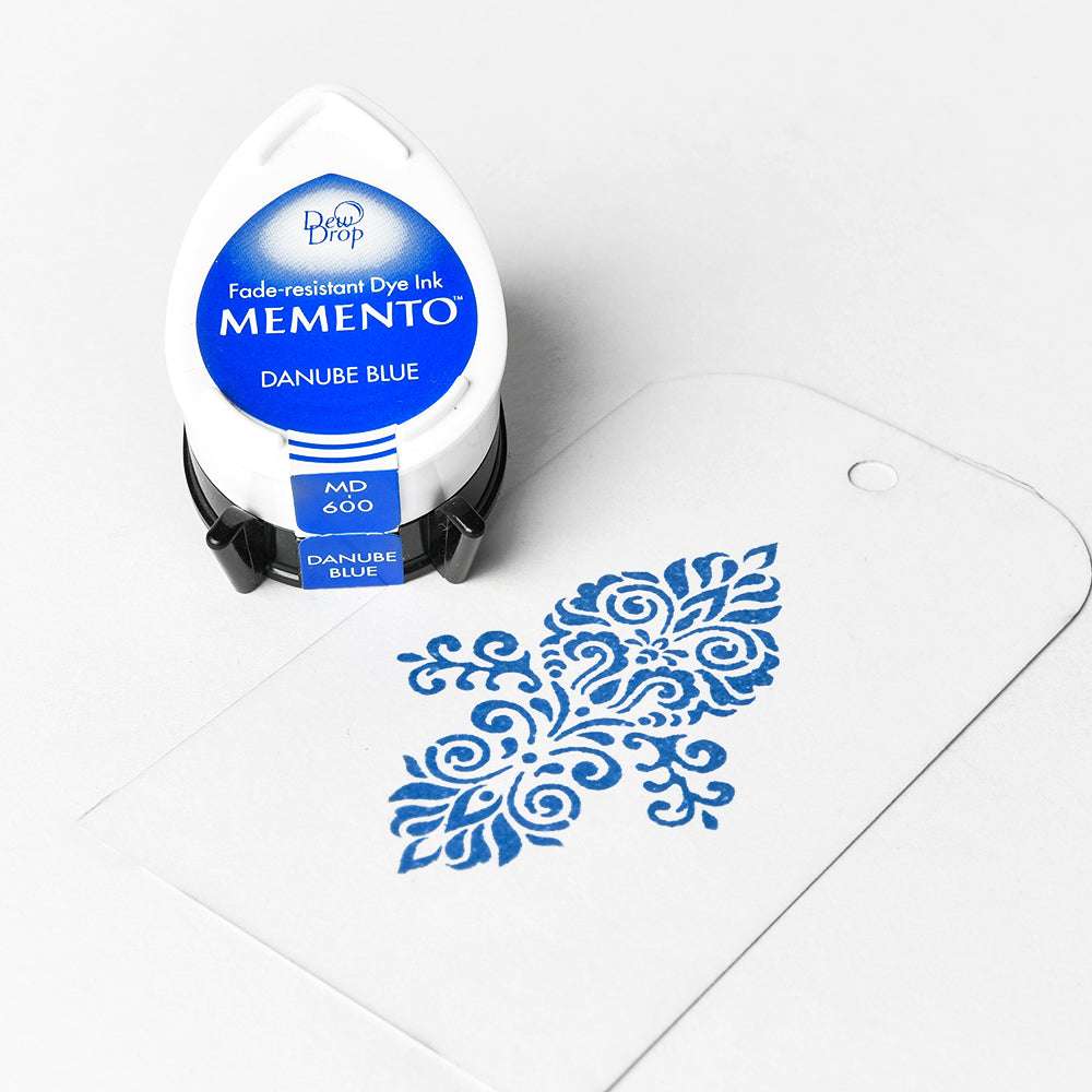 Ein Tsukineko Memento Stempelkissen in Donaublau sitzt neben einem weißen Anhänger mit blauem Blumenmuster - perfekt für wasserbasierte Tinte und detailreiche Stempelmotive.