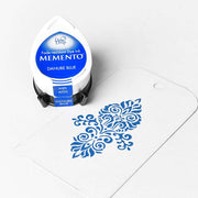 Ein Tsukineko Memento Stempelkissen in Donaublau sitzt neben einem weißen Anhänger mit blauem Blumenmuster - perfekt für wasserbasierte Tinte und detailreiche Stempelmotive.