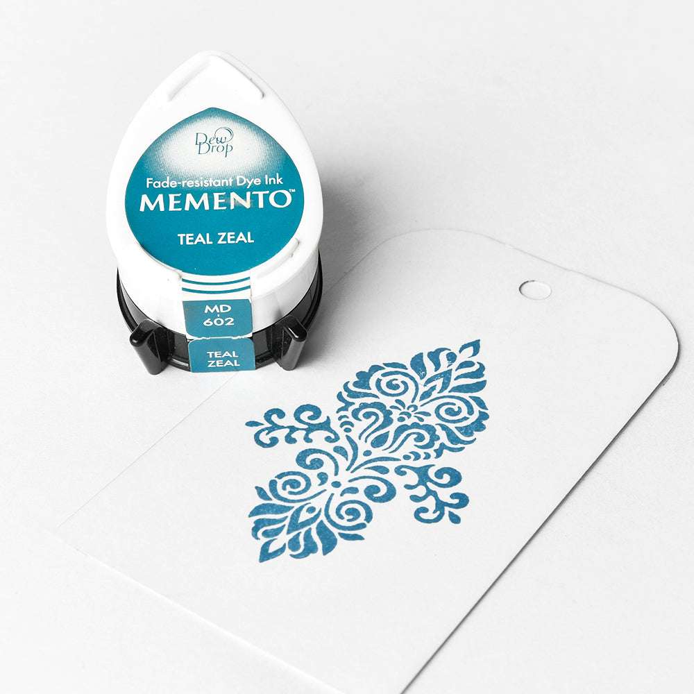 Das tropfenförmige Stempelkissen von Tsukineko mit der Aufschrift Memento Teal Zeal sitzt neben einer weißen Karte mit einem tealfarbenen Dekorationsmuster, das die leuchtenden Farben der Memento Stempelkissen-Linie hervorhebt.