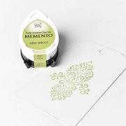 Ein Tsukineko Memento Stempelkissen "New Sprout" mit wasserbasierter Tinte liegt neben einem weißen Tag mit grünem Dekormuster.
