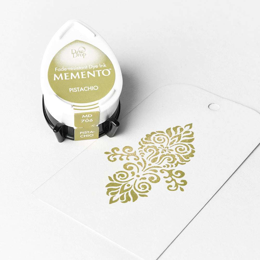 Ein pistaziengrünes Tsukineko Stempelkissen neben einem weißen Anhänger mit einem detailreichen Stempelmotiv in passender Tinte gestempelt.